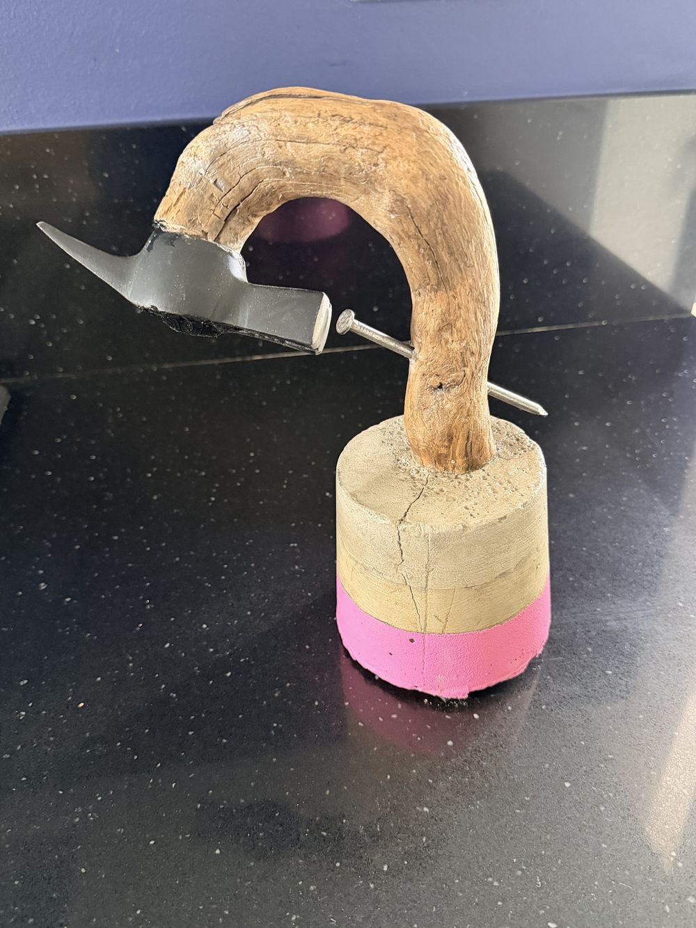 Un ramo curvo con la testa di un martello in cima, che pianta un chiodo nel ramo stesso, su una base di cemento con una fascia rosa