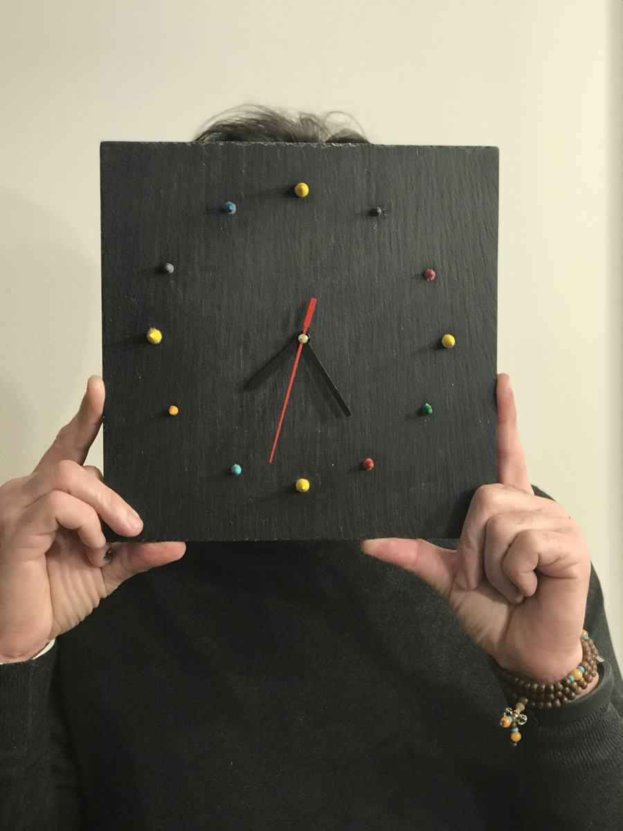 Orologio su ardesia con ore segnate da punti colorati, tenuto davanti al volto