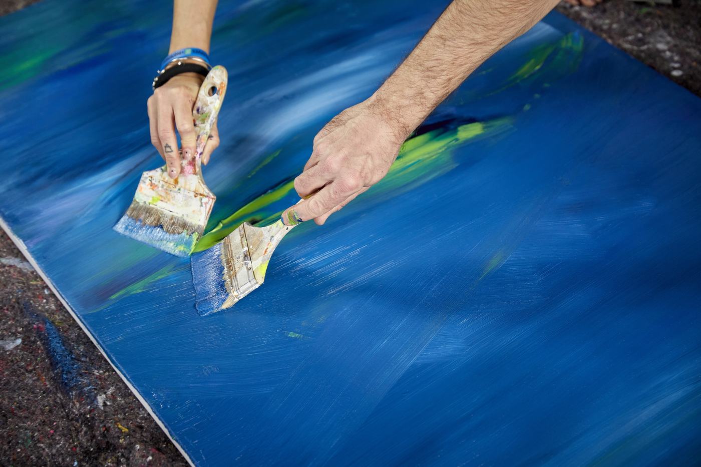 Due mani con pennelli larghi stendono blu e verde sulla tela